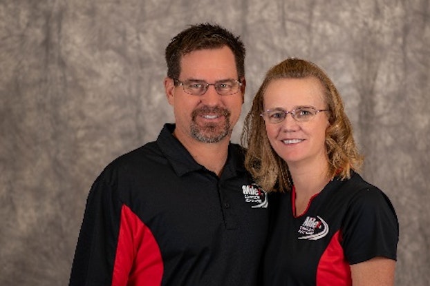  Melissa Riedesel and Grant Humston, Owners of Milex Complete Auto Care, standing against a gray background and wearing matching black-and-red shirts with the Milex logo over the right side of the chest.